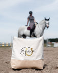 Helle Jutetasche mit schwarz-goldenem Motiv auf einem Reitplatz vor weißem Pferd