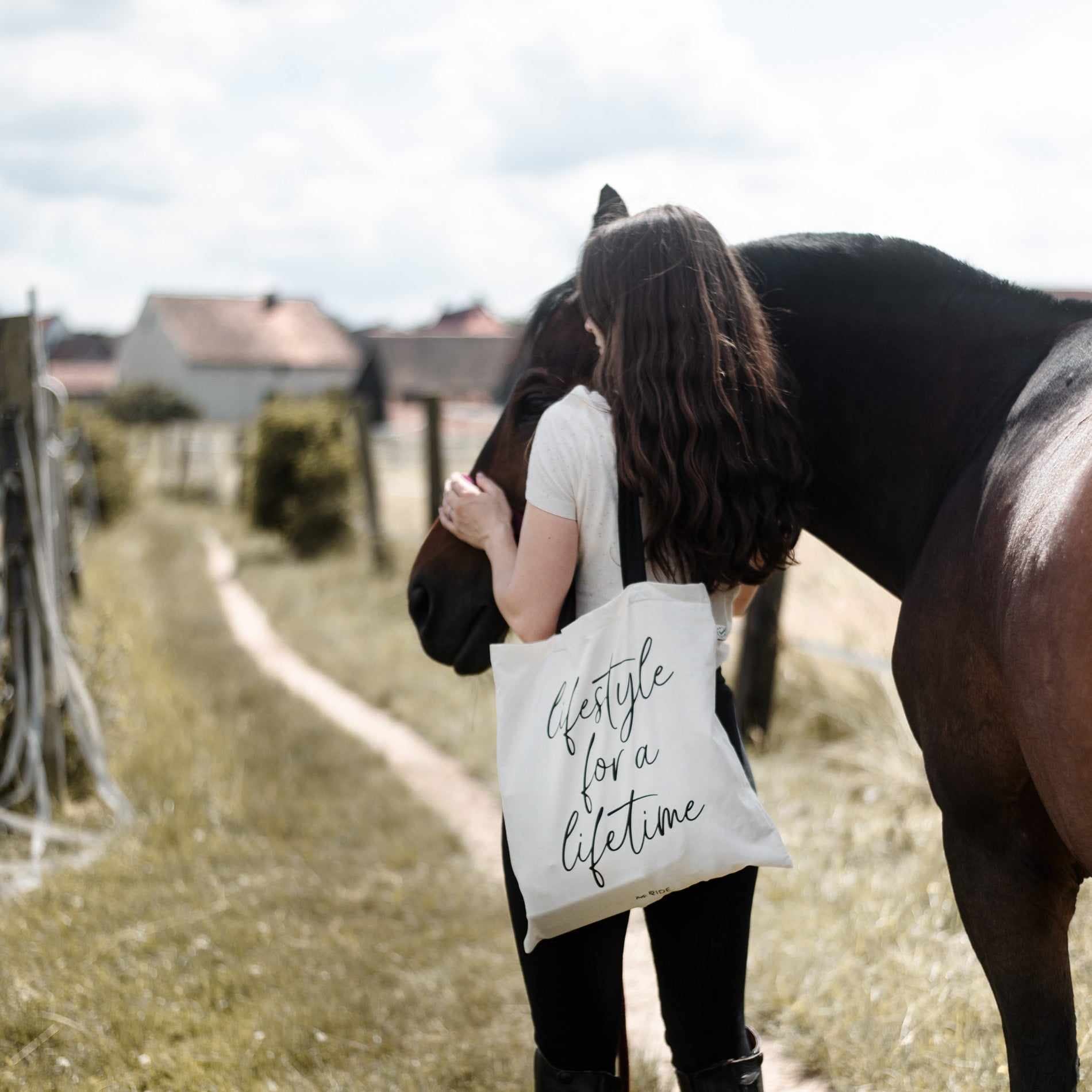 Junge Frau mit heller Baumwolltasche neben einem braunen Pferd auf einem Feldweg