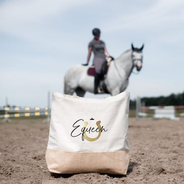 Helle Jutetasche mit schwarz-goldenem Motiv auf einem Reitplatz vor weißem Pferd